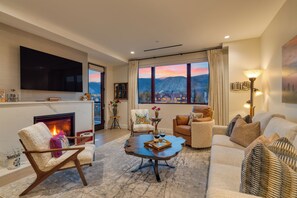 A living room with a fireplace, a television, a large window with a mountain view, and a cozy seating area. - Condo + Ski Shuttle + Pool + Spa Access | Mountain Dream at Frontgate (Avon)
