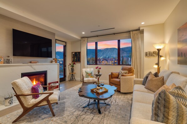 A living room with a fireplace, a television, a large window with a mountain view, and a cozy seating area. - Condo + Ski Shuttle + Pool + Spa Access | Mountain Dream at Frontgate (Avon)