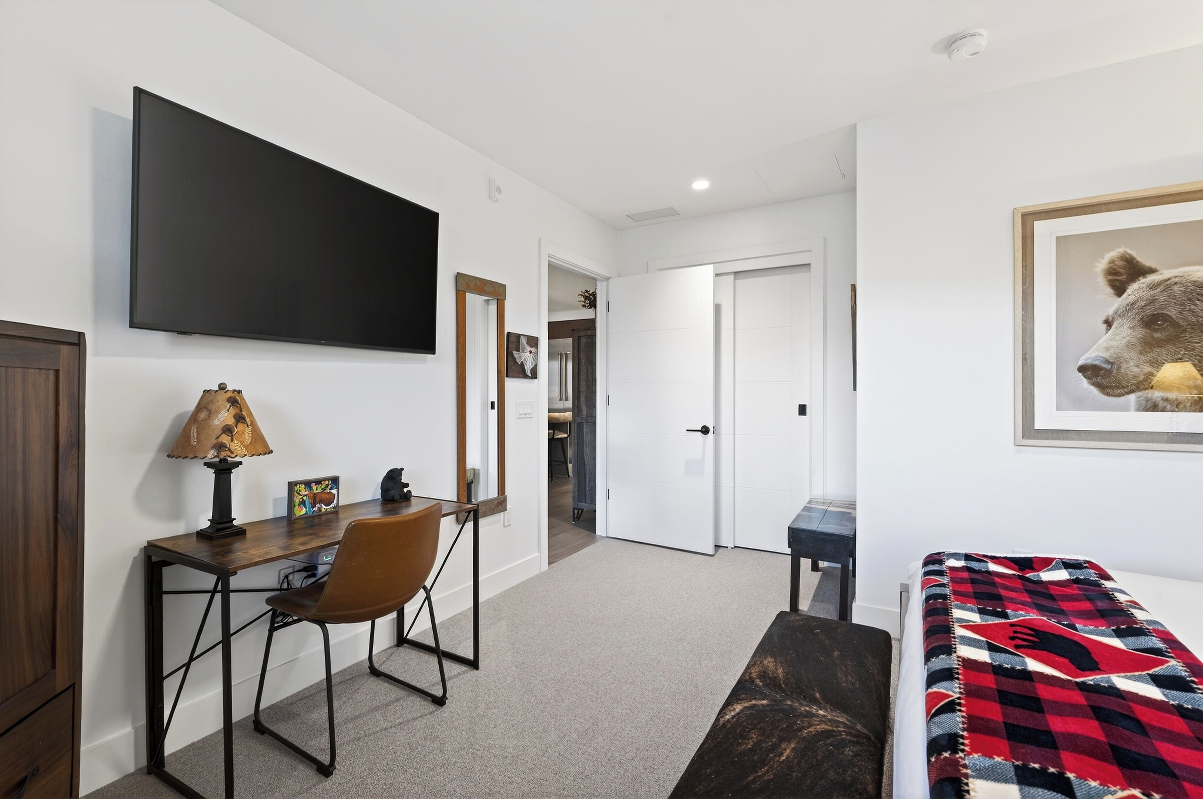 A bedroom with a bed, a desk with a chair, a television, and a door leading to another room.