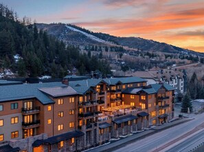 A multi-story hotel with balconies and large windows, set against a backdrop of snow-covered hills and a sunset sky. - Condo + Ski Shuttle + Pool + Spa Access | Mountain Dream at Frontgate (Avon)
