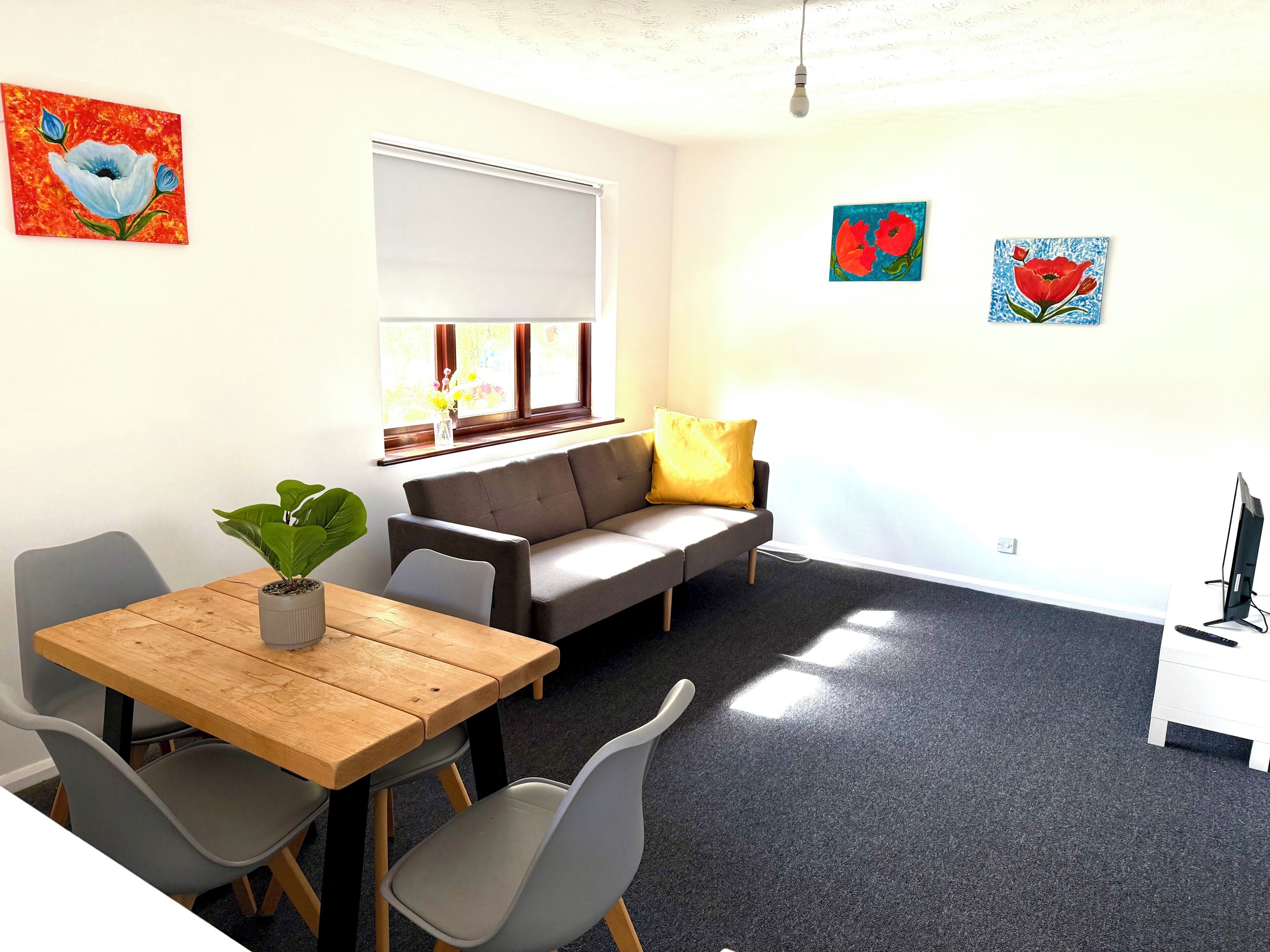 A room with a wooden table, grey chairs, a grey sofa with a yellow pillow, a white desk with a computer, and artwork on the walls.