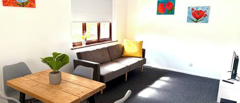A room with a wooden table, grey chairs, a grey sofa with a yellow pillow, a white desk with a computer, and artwork on the walls.
