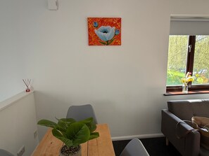 A room with a wooden table, a potted plant, a gray couch, a window with a view of trees, and a painting of flowers on the wall. - Private Maisonette Chandlers Ford (Chandler's Ford)