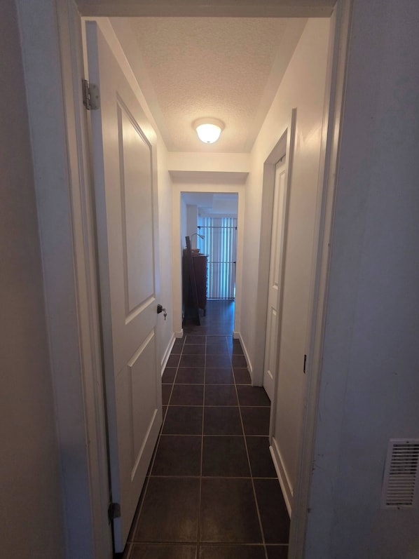A narrow hallway with white walls and dark tiled flooring, leading to a room with a window and blinds.