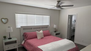 Ein Schlafzimmer mit einem Bett, zwei Nachttischen, einem Deckenventilator, einem Fenster mit Jalousien und einem Spiegel an der Wand.