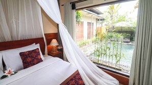 Uma cama com almofadas de padrão branco e vermelho, uma luminária de cabeceira e vista para uma piscina e vegetação lá fora.