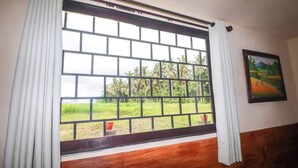 Quarto com uma grande janela com grade de metal preta, cortinas brancas e vista para uma paisagem verde com árvores e um campo.