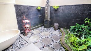 Een klein, afgesloten buitenruimte met een vloer van stenen tegels, een groot wit wasbak, een metalen plank met flessen, en potplanten langs de muren.