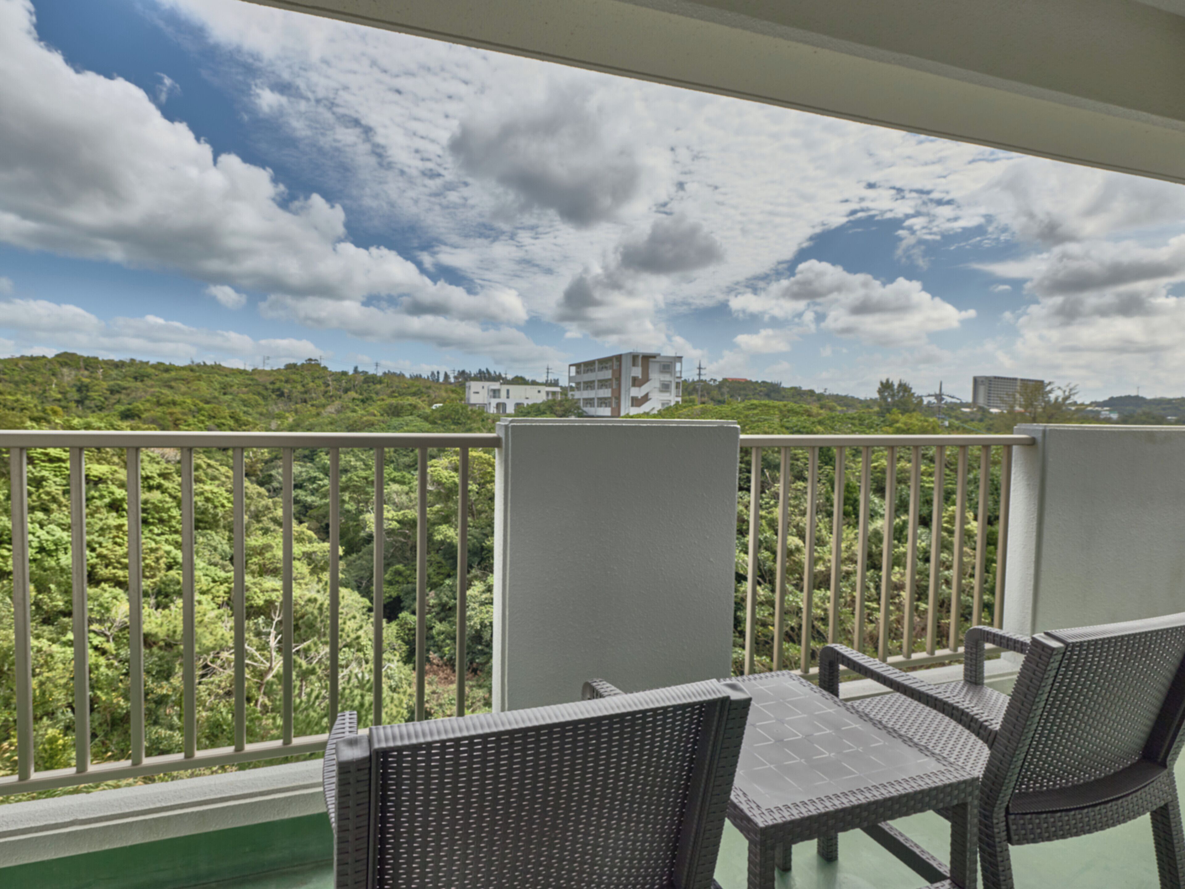 A balcony with wicker chairs, a view of a green hillside, and buildings in the distance.