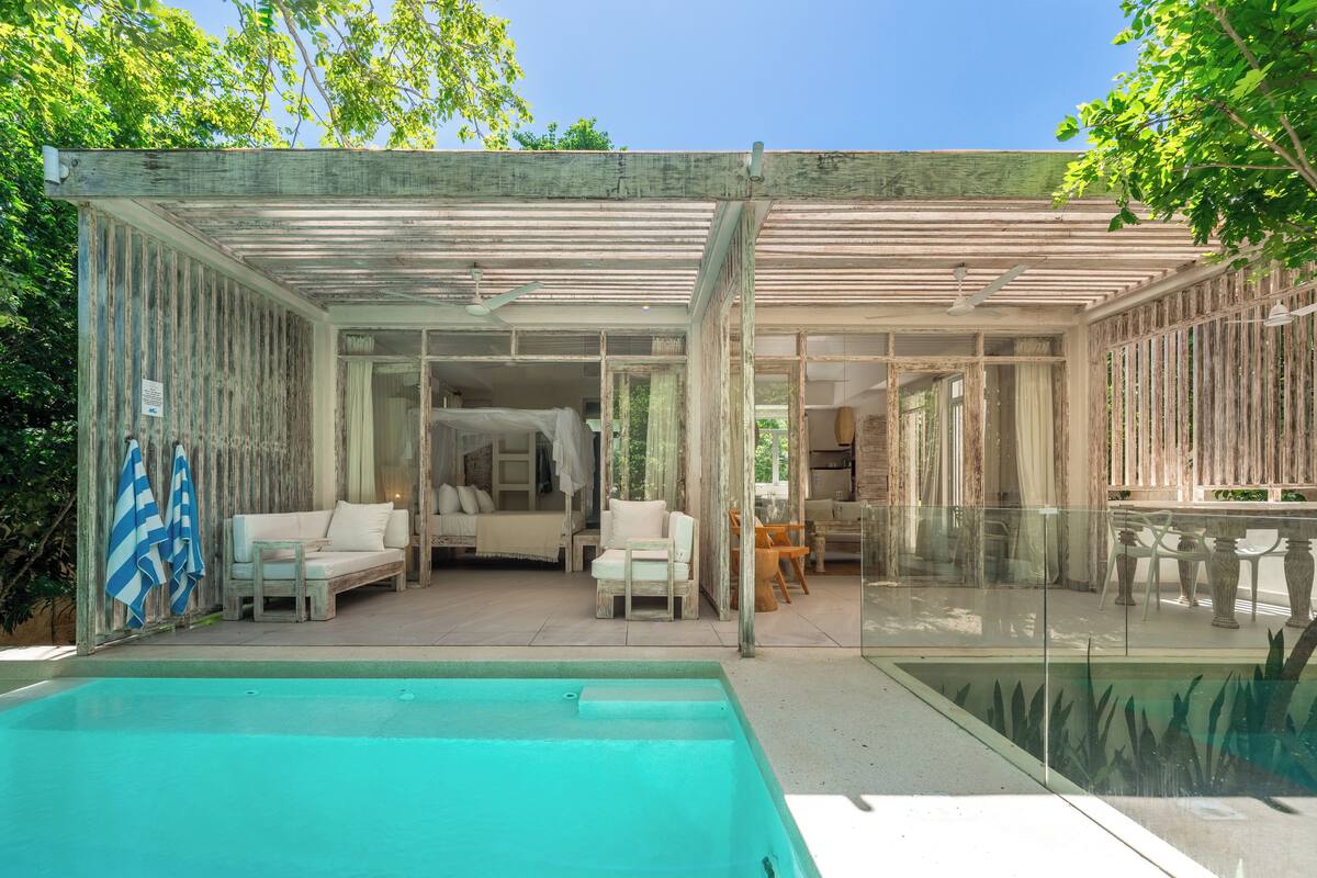 A modern outdoor pool area with a glass enclosure, white furniture, and a view of the interior.