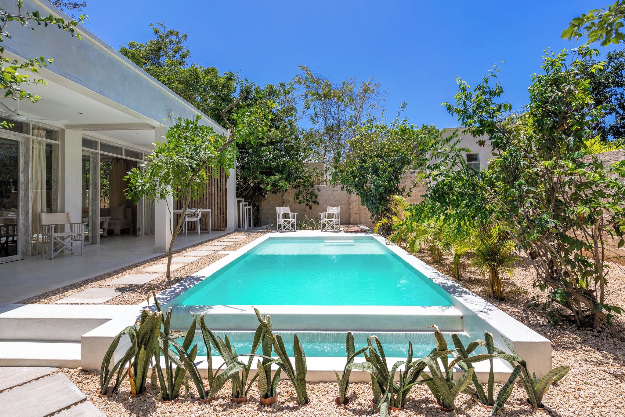 A swimming pool with a white deck, surrounded by small plants and trees, and a patio area with outdoor furniture.