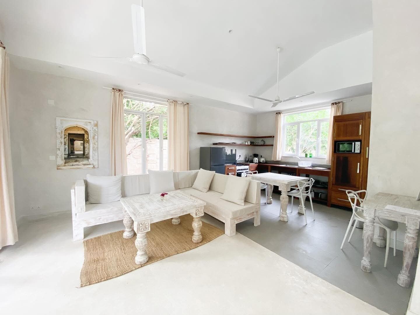 A spacious living area with a sofa, coffee table, and dining table, a kitchen with appliances, and large windows.
