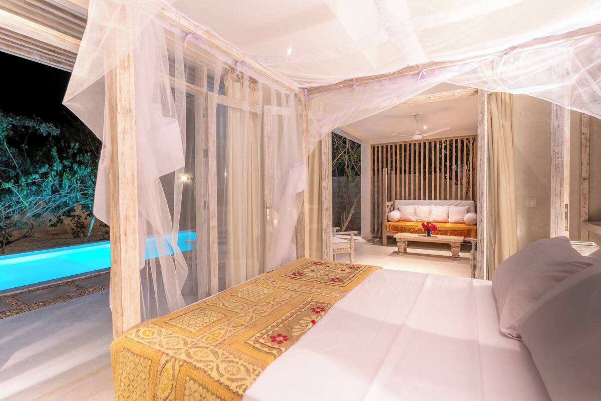 A bedroom with a large bed, a bedside table, and a bench. There is a window with sheer curtains, and a view of the outside.