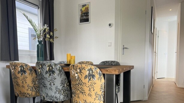 Een eetgedeelte met een houten tafel en vier stoelen met bloemenmotieven. Op de vensterbank staat een vaas met bloemen, en aan de muur hangt een lijst met een afbeelding.