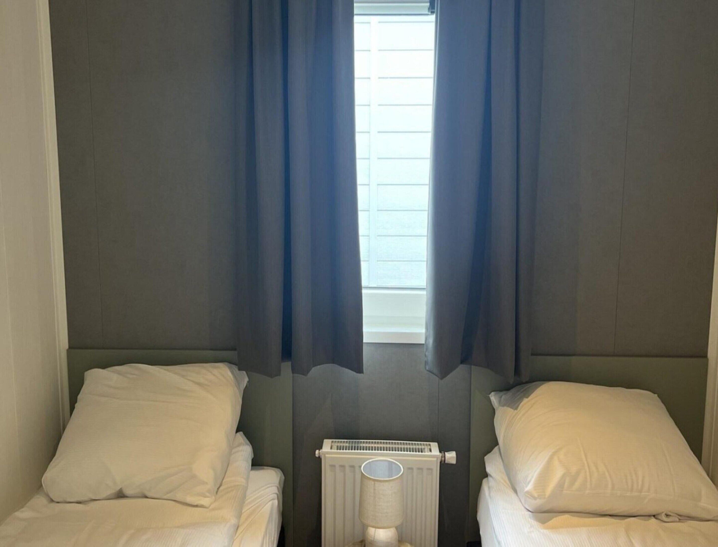 A small, dimly lit room with two single beds, a radiator, and a window with blue curtains.