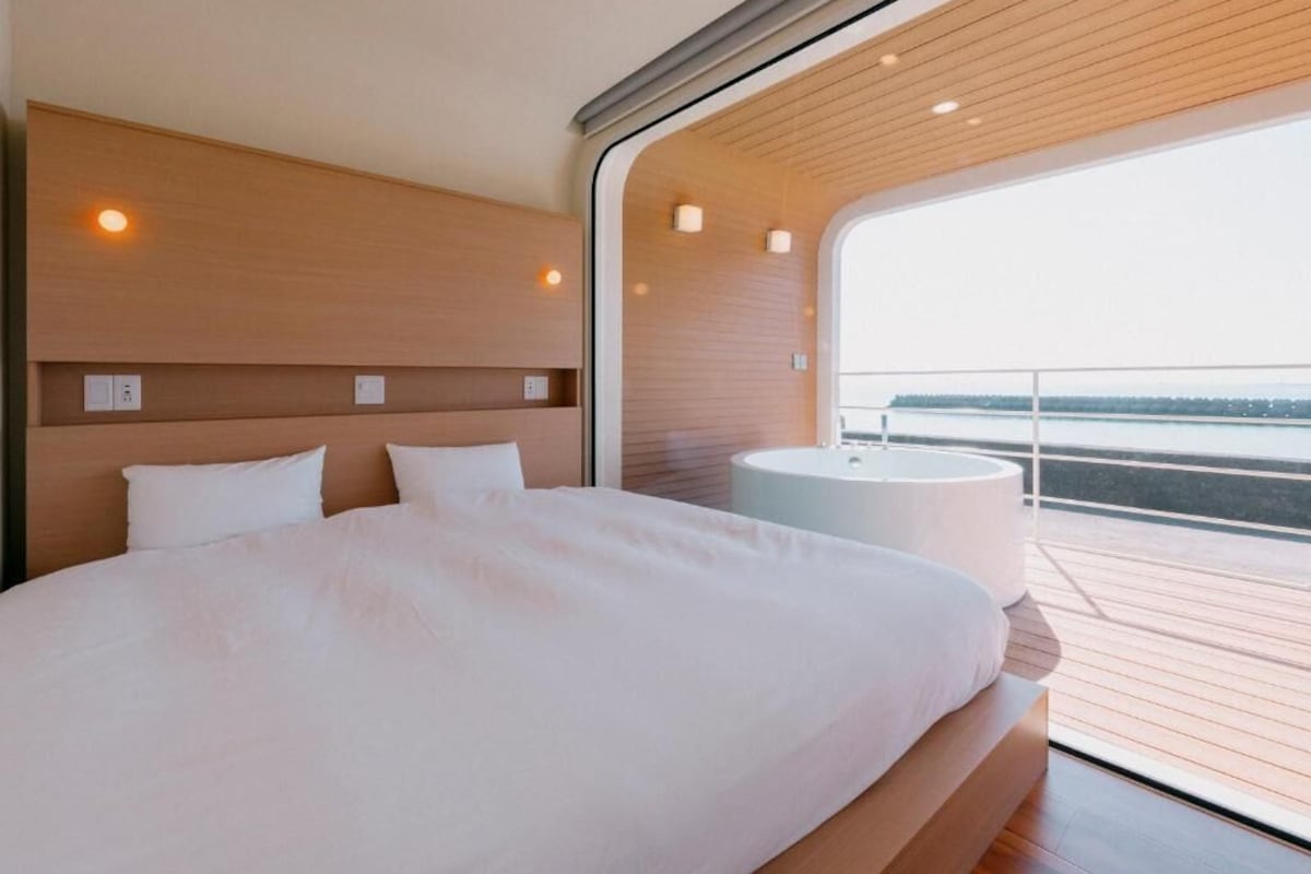 A modern bedroom with a large bed, a round bathtub, and a view of the water.
