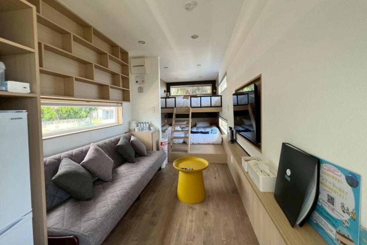 A compact room with a sofa, a yellow stool, bunk beds, and a desk with a computer.