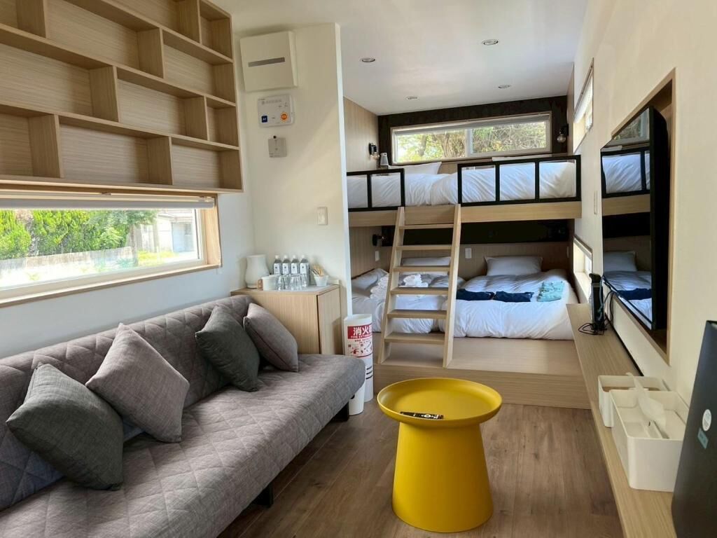 Una habitación pequeña con literas, un sofá, un taburete amarillo y una ventana.