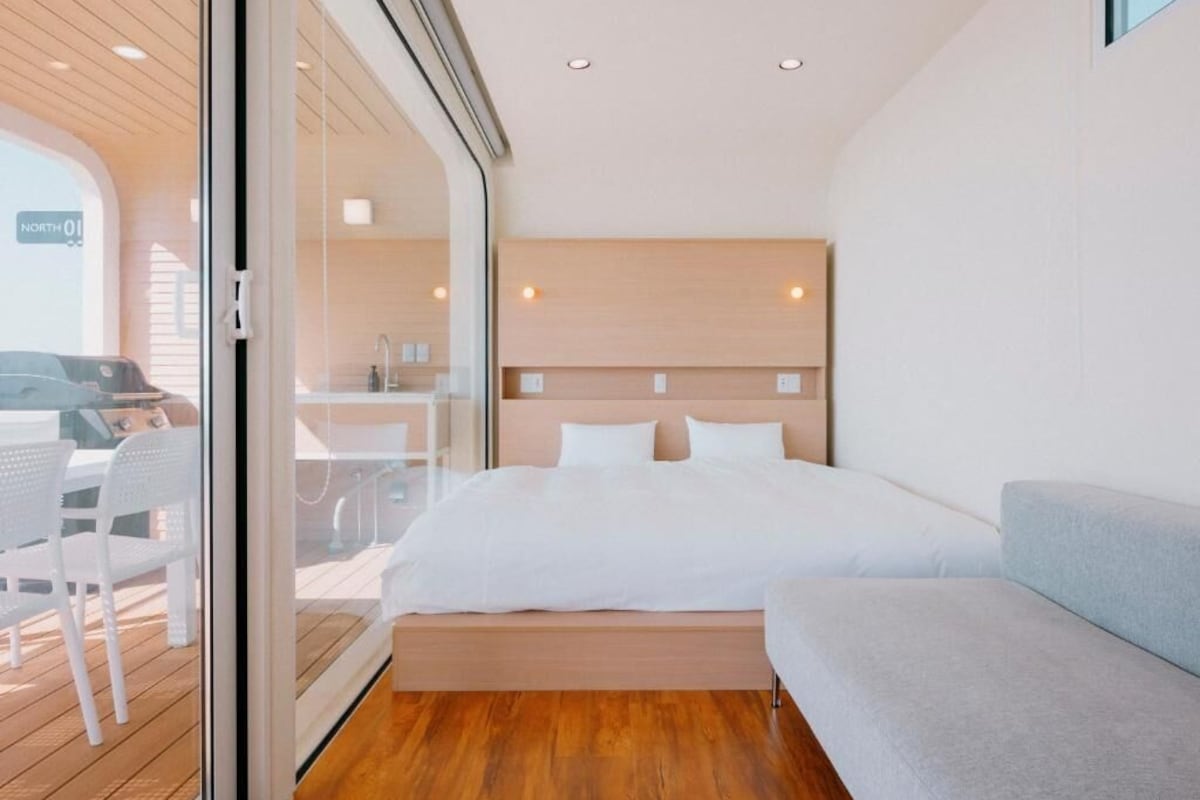 A modern hotel room with a bed, a sofa, and a glass door leading to an outdoor area.