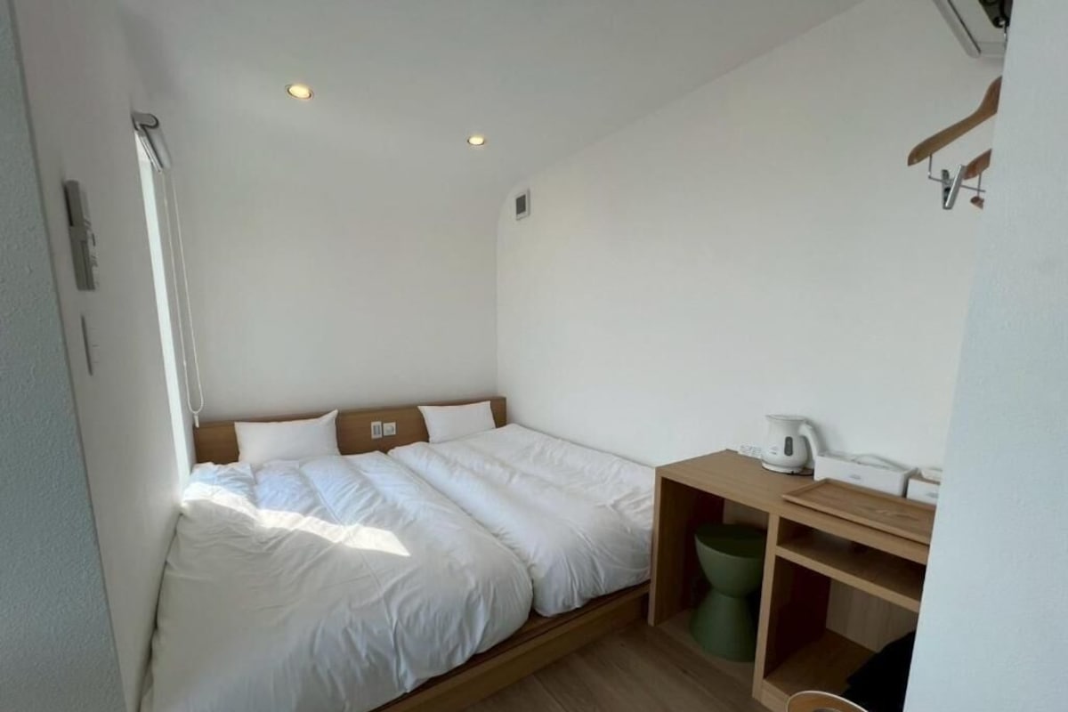 A small, single-bed room with a wooden headboard, a bedside table with a kettle, and a green trash bin.
