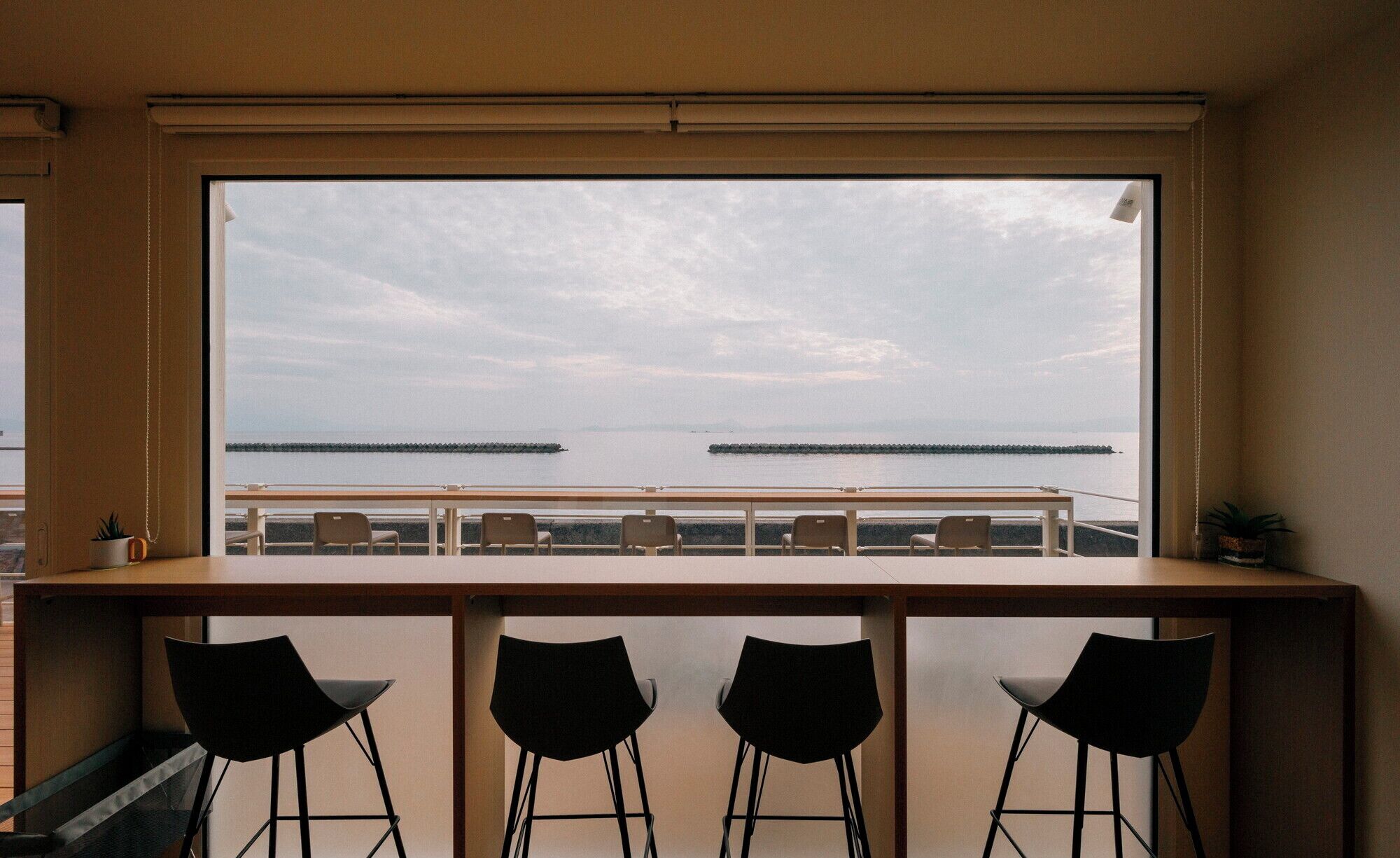 A room with a large window overlooking a body of water and a pier. There are three black chairs and a wooden table in the foreground.