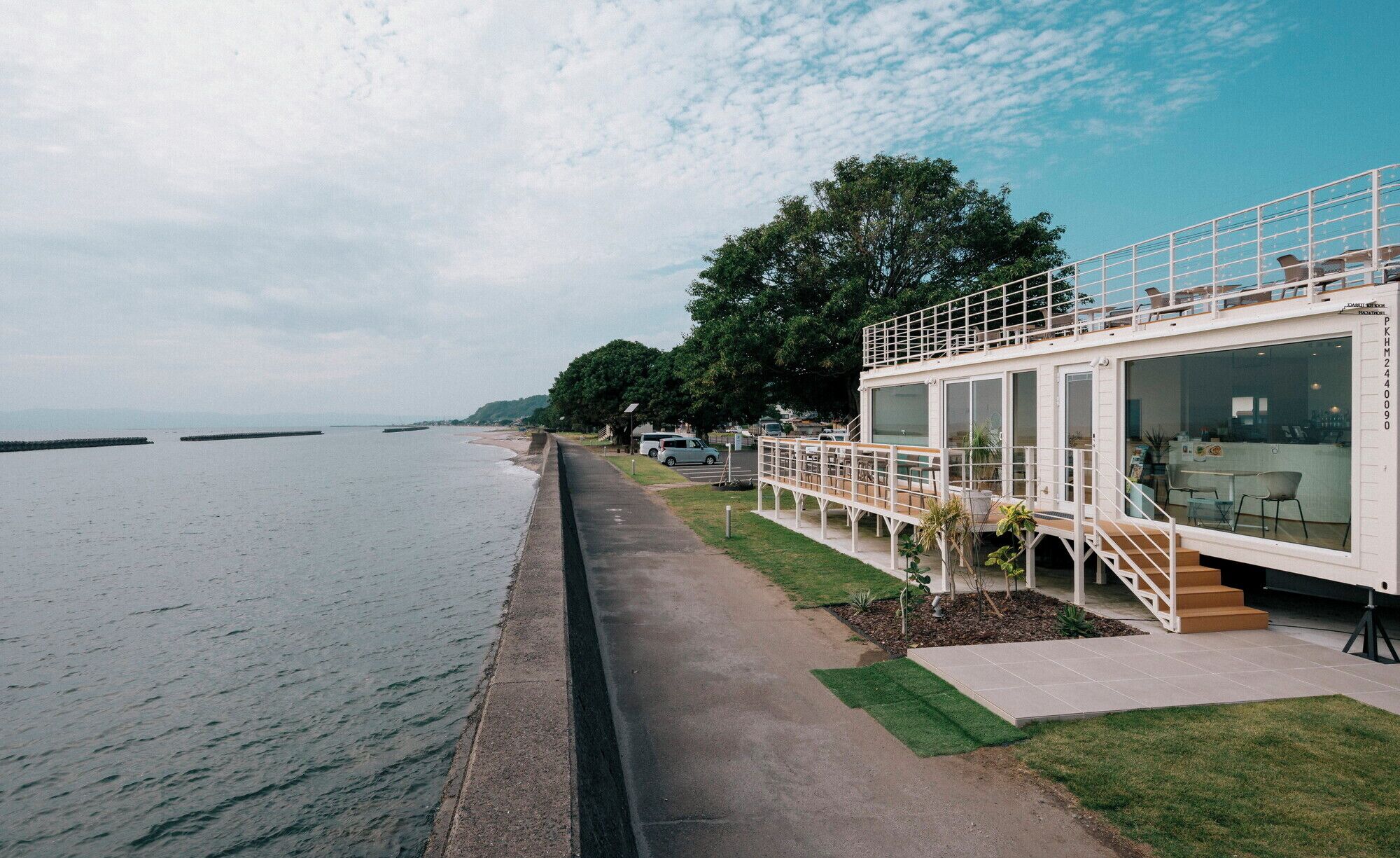 A coastal building with a deck overlooking the sea, a paved walkway, and a grassy area with small plants.