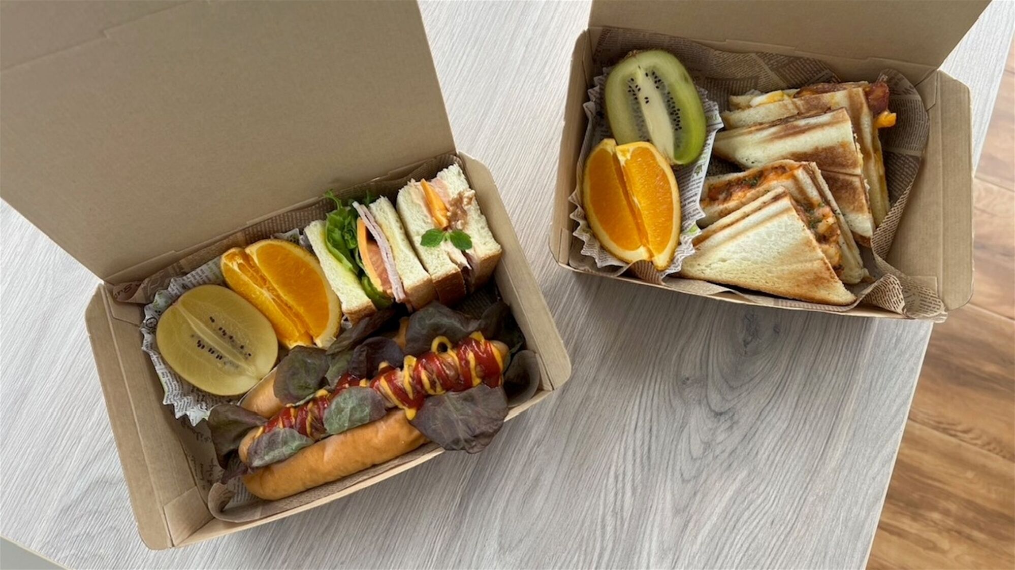 Dos cajas de cartón abiertas con sándwiches y frutas cortadas sobre una superficie de madera.