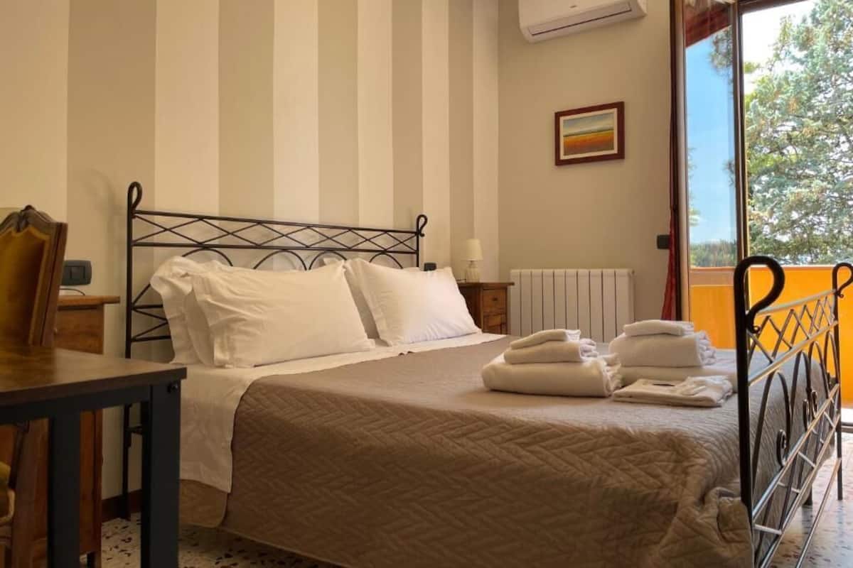Un lit bien fait, avec du linge de lit blanc, une table de chevet avec une lampe et une fenêtre donnant sur des arbres.