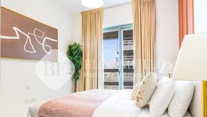 A modern bedroom with a large bed, beige curtains, a view through a glass door, and a wall-mounted abstract artwork. - Bloomfields 2br Unique Ajwan Soul B (Abu Dhabi)