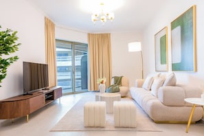A modern living room with a sofa, ottomans, a coffee table, a TV, and a large window with curtains. - Bloomfields 2br+1 in Ajwan Towers (Abu Dhabi)