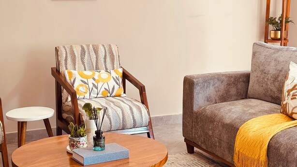 A living room with a round wooden coffee table, a grey sofa with a yellow throw, a striped armchair, and a small white side table. - Liora Haven - 2BR Home That Feels Just Right (Arusha)
