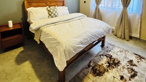 A bedroom with a wooden bed, a nightstand, a framed picture on the wall, and a window with curtains. - Liora Haven - 2BR Home That Feels Just Right (Arusha)