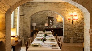 Une salle à manger aux murs de pierre, avec une longue table dressée pour un repas, flanquée de lampes à bougies et d’un lustre.