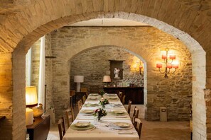 Ein Esszimmer mit steinernen Wänden, in dem ein langer Tisch für eine Mahlzeit gedeckt ist, flankiert von kerzenbeleuchteten Lampen und einer Kristallleuchter.