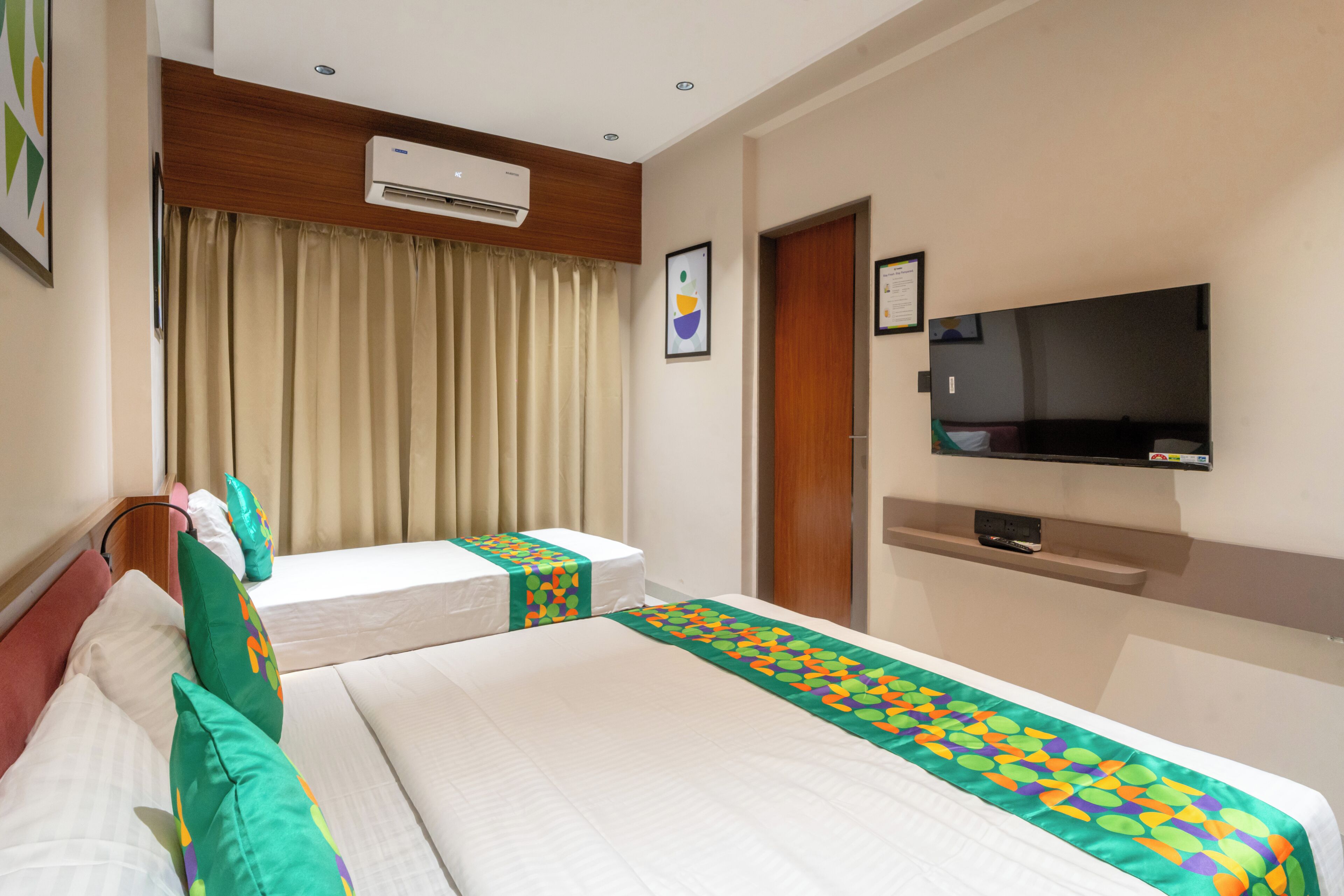 A hotel room with two beds, a TV, an air conditioner, and a window with curtains.