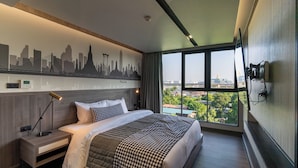 Une chambre d’hôtel moderne avec un grand lit, une fresque murale représentant le paysage urbain, une table de chevet avec une lampe et une vue sur la ville à travers de grandes fenêtres.