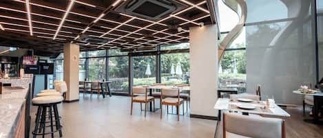 A modern restaurant interior with a grid ceiling, large windows, and a mix of bar and dining areas.