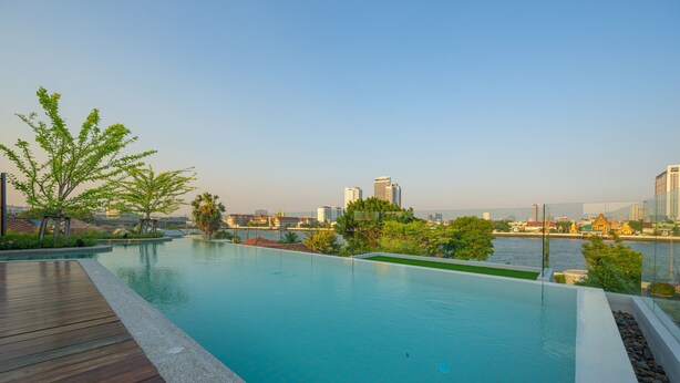A rooftop infinity pool with a wooden deck, surrounded by greenery and overlooking a cityscape with modern buildings and a river. - We Hotel Riverfront (Bangkok)
