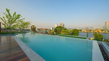 A rooftop infinity pool with a wooden deck, surrounded by greenery and overlooking a cityscape with modern buildings and a river.