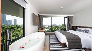 A modern hotel room with a large bathtub, a bed with a grey blanket, a small table with a fruit bowl, and a city view.