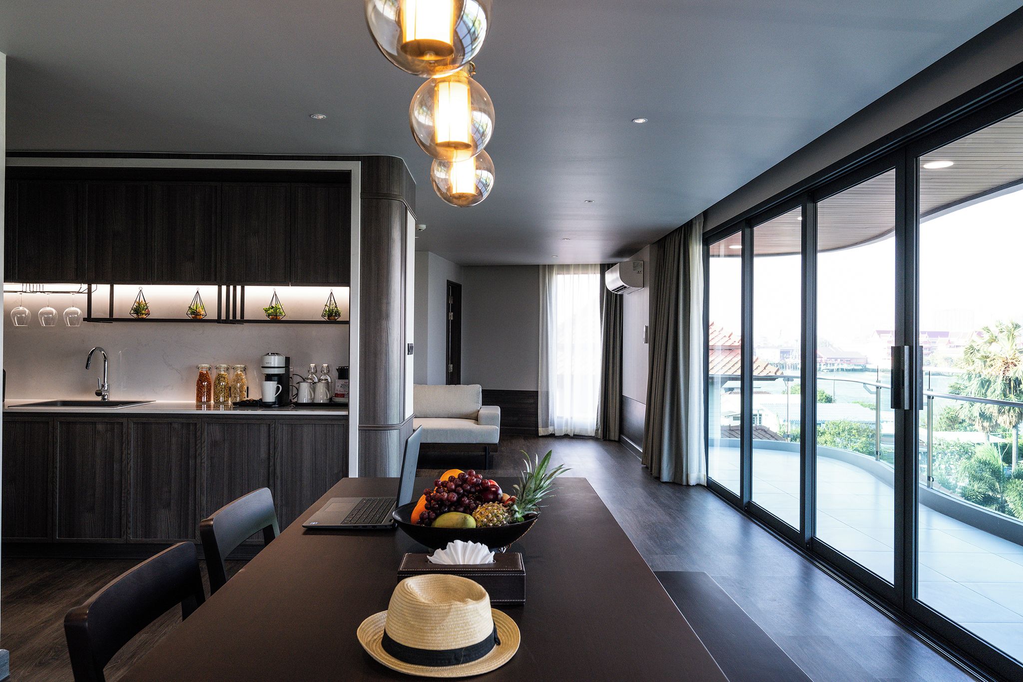 A modern kitchen with dark wood cabinets, a dining area with a large table, and a view through large windows.