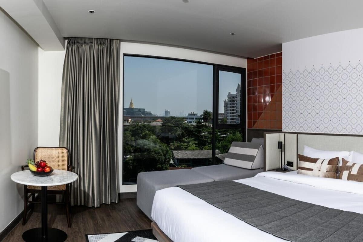 A hotel room with a bed, a small table with fruit, a view of the city, and a large window with curtains.