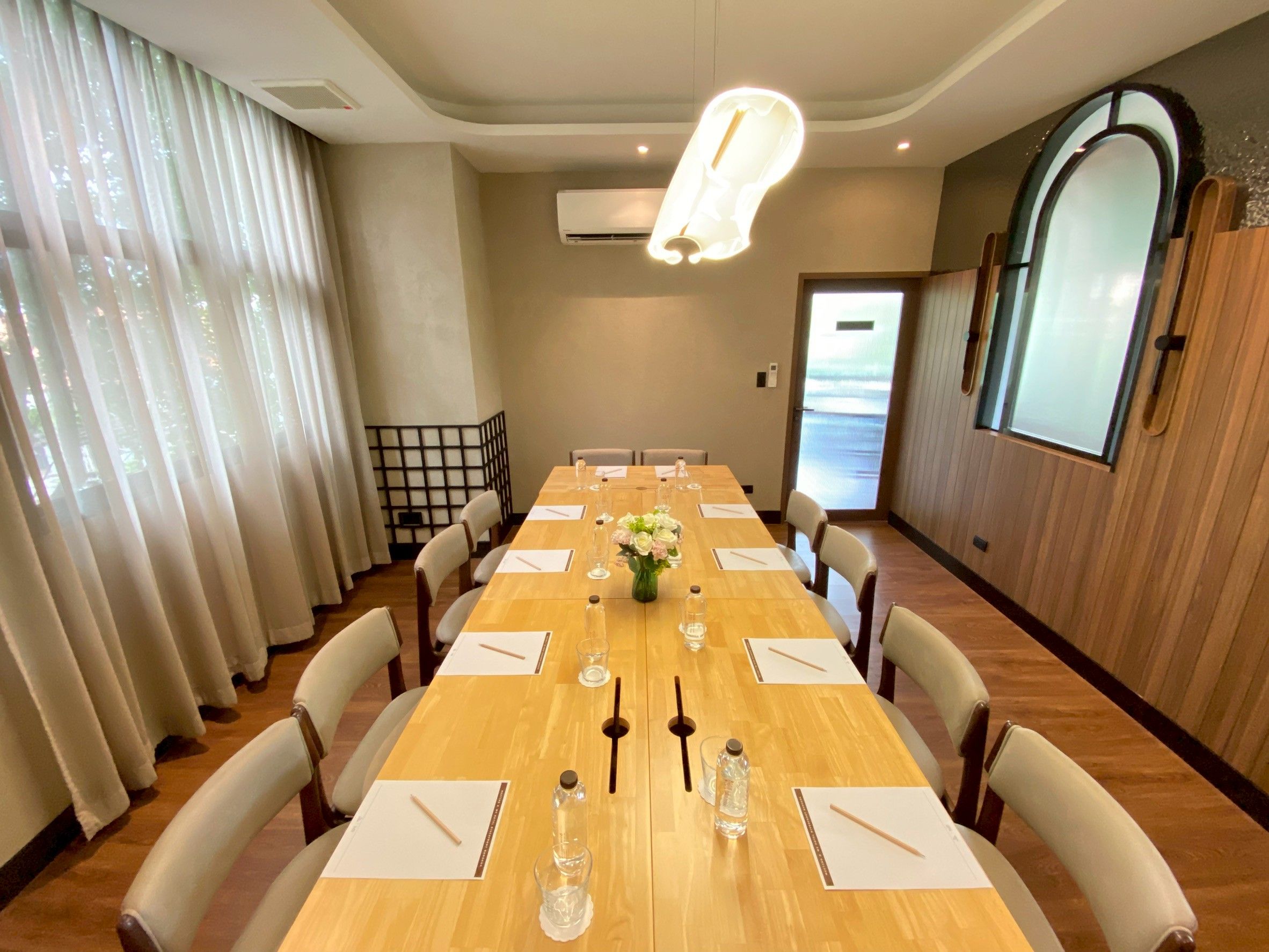 A conference room with a long table, chairs, a vase with flowers, and a large window with curtains.