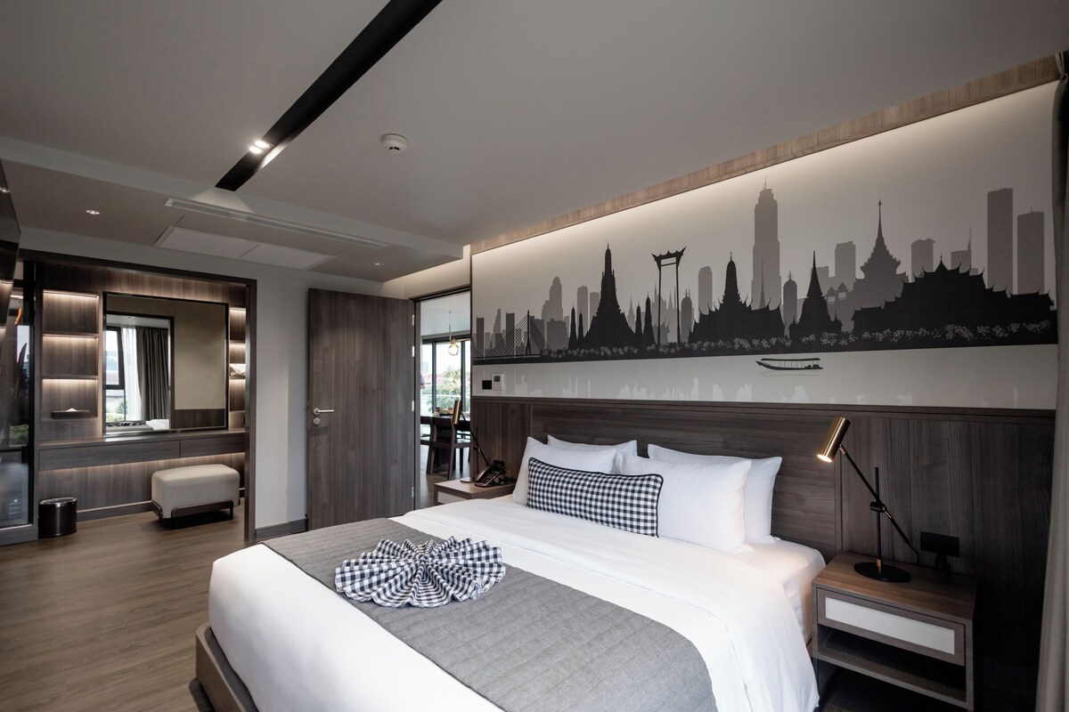 A modern hotel room with a large bed, a city skyline mural, a wooden headboard, and a nightstand with a lamp.