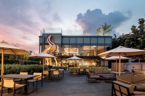 An outdoor patio area with tables and chairs, umbrellas, and a modern building with a glass facade.