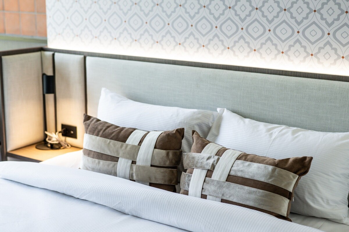Un letto rifatto con cura, lenzuola bianche e due cuscini decorativi con motivo intrecciato.
