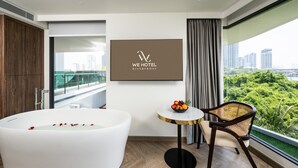 A modern hotel room with a large bathtub, a dining table with fruit, a chair, a city view, and a framed logo on the wall. - We Hotel Riverfront (Bangkok)