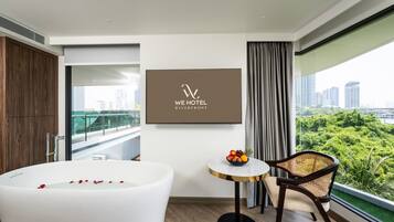 A modern hotel room with a large bathtub, a dining table with fruit, a chair, a city view, and a framed logo on the wall.