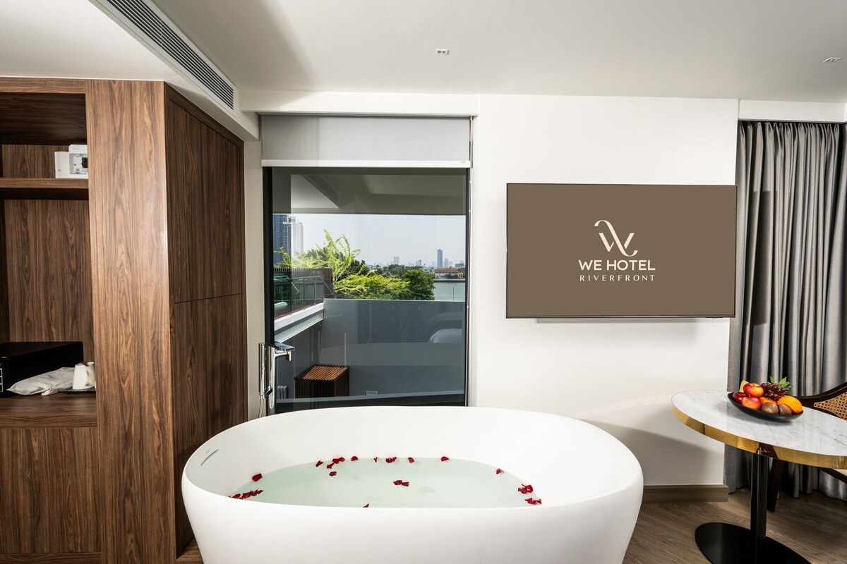Une chambre d’hôtel moderne dotée d’une grande baignoire, d’un balcon offrant une vue sur la ville et d’un coin repas agrémenté d’un panier de fruits.