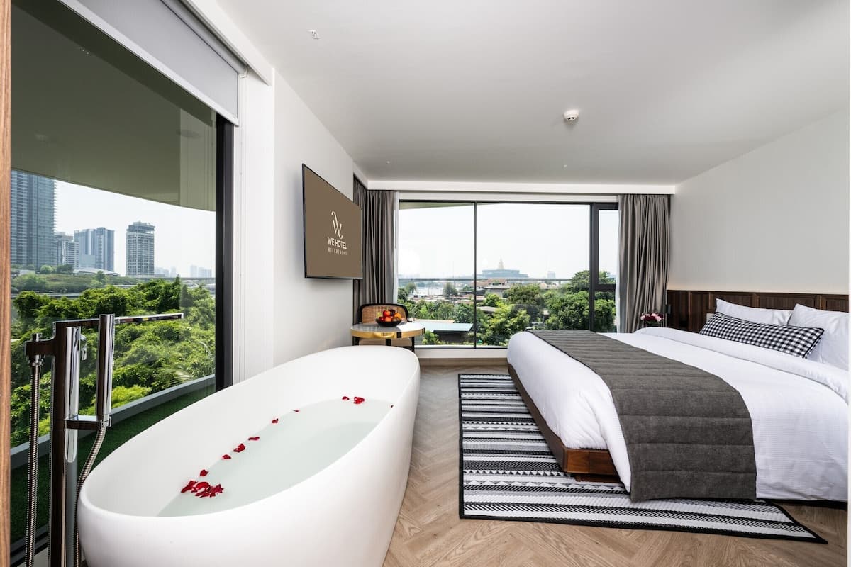 A modern hotel room with a large bed, a bathtub with rose petals, a city view, and a small table with fruit.